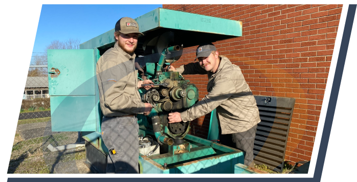 Two people work on an older generator unit painted in bright teal against a brick wall